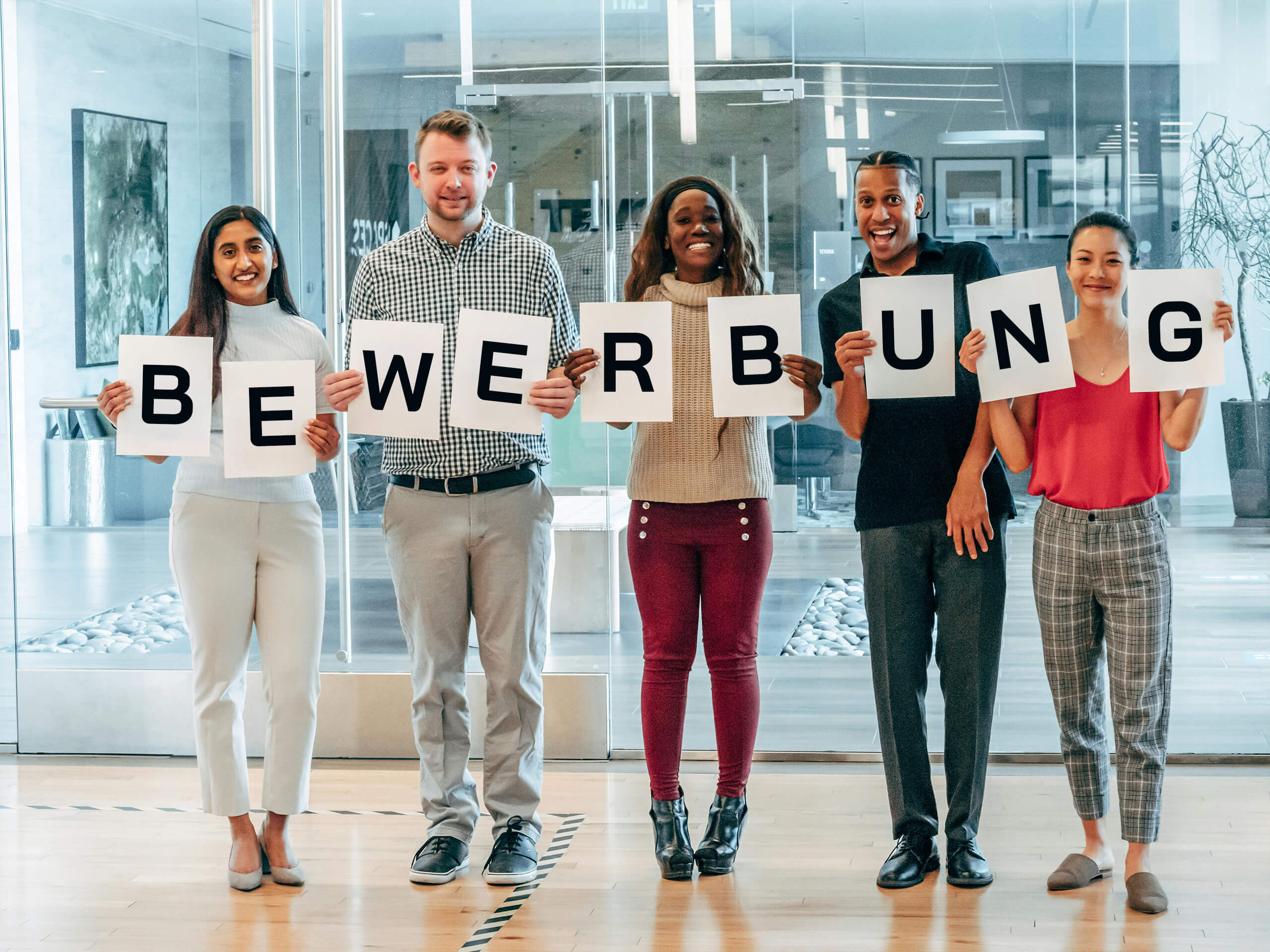 Lächelnde diverse Menschen halten ein weißes Blatt in der Hand, auf dem ein schwarzer Buchstabe steht. Daraus wird das Wort "Bewerbung" gebildet.